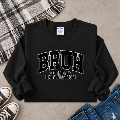 A black crewneck sweatshirt with 'BRUH -FORMERLY KNOWN AS MOM' printed in white on the front, styled in a flat lay with jeans, sneakers, a plaid blanket, and a gray textured surface.
