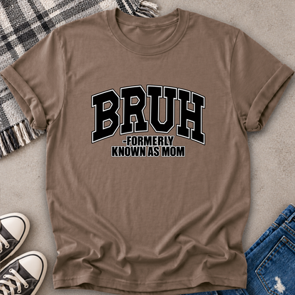 A brown short-sleeve graphic T-shirt laid flat on a textured surface, with black-and-white text reading “BRUH -FORMERLY KNOWN AS MOM.” Styled flat-lay props include a plaid blanket, jeans, sneakers, and a neutral surface.