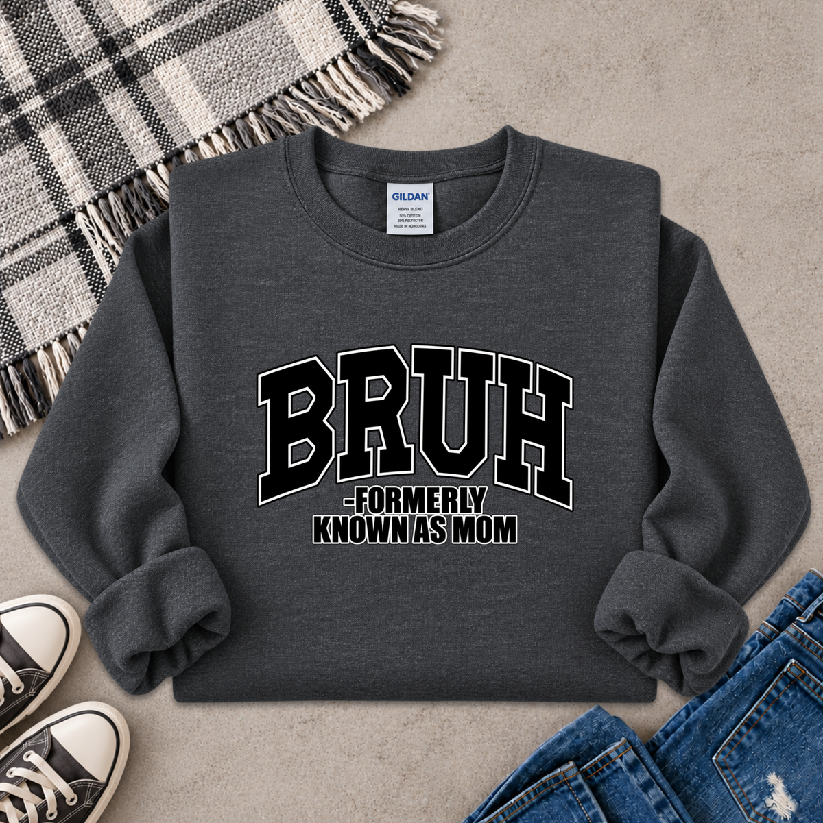 A dark gray crewneck sweatshirt with 'BRUH -FORMERLY KNOWN AS MOM' printed in black and white varsity lettering, styled in a flat lay with jeans, sneakers, a plaid blanket, and a concrete floor background.