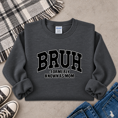 A dark gray crewneck sweatshirt with 'BRUH -FORMERLY KNOWN AS MOM' printed in black and white varsity lettering, styled in a flat lay with jeans, sneakers, a plaid blanket, and a concrete floor background.