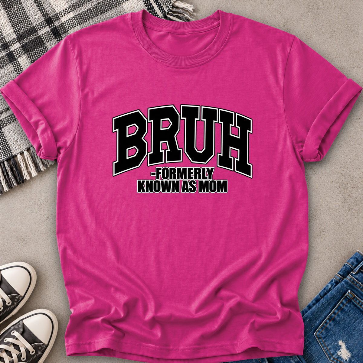 A bright pink short-sleeve t-shirt with a black and white graphic reading 'BRUH -FORMERLY KNOWN AS MOM', styled in a flat lay with jeans, sneakers, a plaid blanket, and a gray textured surface.