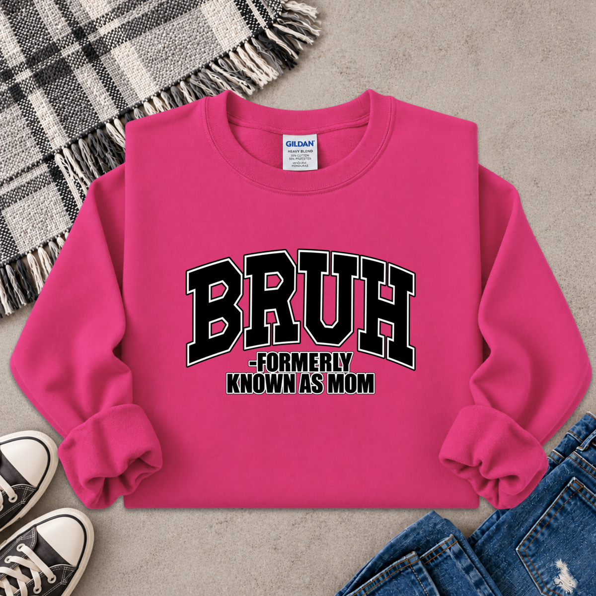 A bright pink crewneck sweatshirt laid flat on a gray surface, featuring the text 'BRUH -FORMERLY KNOWN AS MOM' in black and white varsity lettering. The sweatshirt is styled with a plaid blanket, black sneakers, and blue jeans.