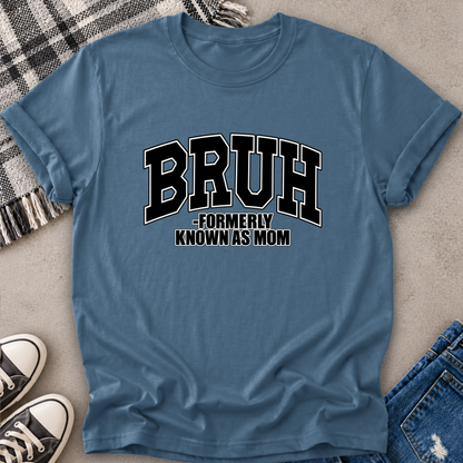 A slate blue graphic t-shirt laid flat on a textured gray surface, styled with a plaid blanket, black sneakers, and blue jeans. The t-shirt features the text 'BRUH -FORMERLY KNOWN AS MOM' in black and white varsity-style lettering.
