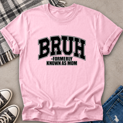 A light pink short-sleeve t-shirt with a black graphic text reading 'BRUH -FORMERLY KNOWN AS MOM', laid flat on a gray surface with jeans, sneakers, and a plaid blanket partially visible around it.