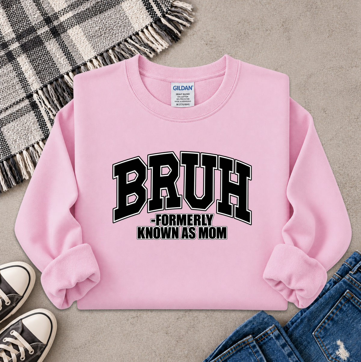 A light pink crewneck sweatshirt with 'BRUH -FORMERLY KNOWN AS MOM' printed in black varsity lettering on the front. The sweatshirt is styled in a flat lay with a plaid blanket, jeans, sneakers, and a gray concrete floor background.
