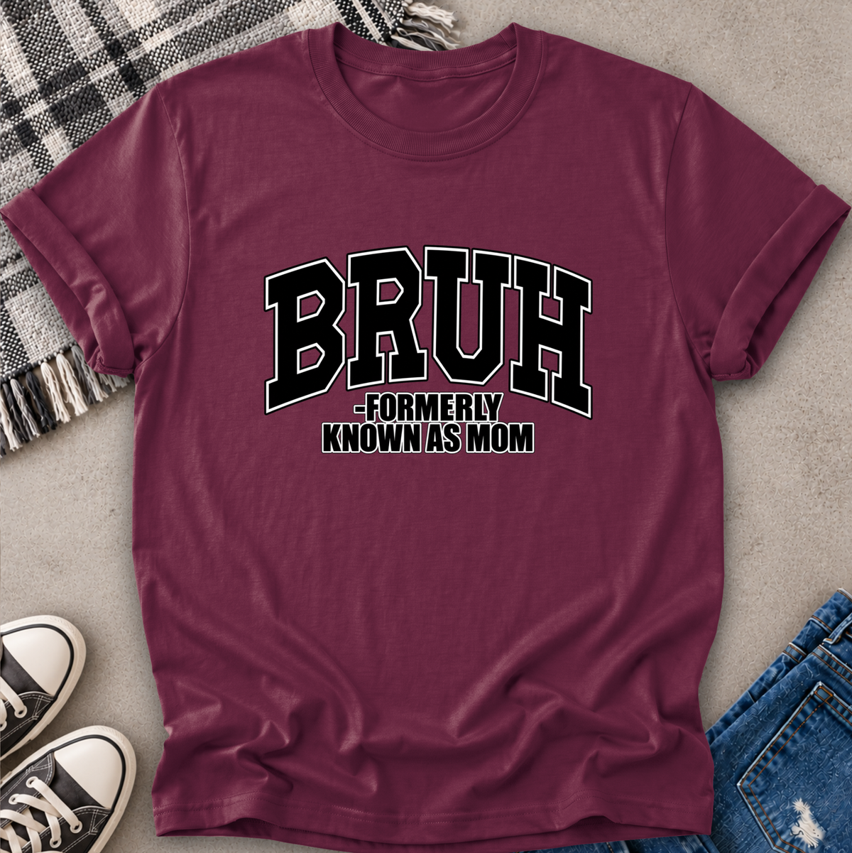 A maroon short-sleeve graphic T-shirt laid flat on a light gray surface, styled with a plaid blanket, black sneakers, and blue jeans. The shirt has large black collegiate-style text reading “BRUH” with smaller text underneath reading “FORMERLY KNOWN AS MOM.