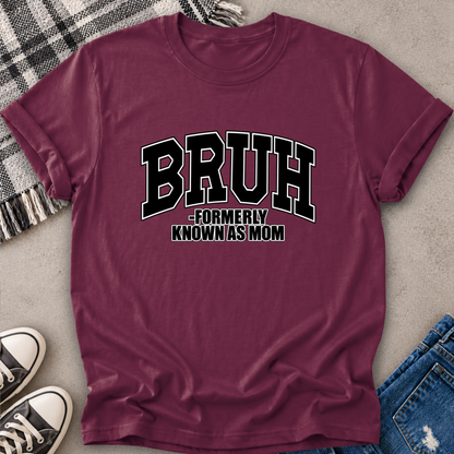 A maroon short-sleeve graphic T-shirt laid flat on a light gray surface, styled with a plaid blanket, black sneakers, and blue jeans. The shirt has large black collegiate-style text reading “BRUH” with smaller text underneath reading “FORMERLY KNOWN AS MOM.