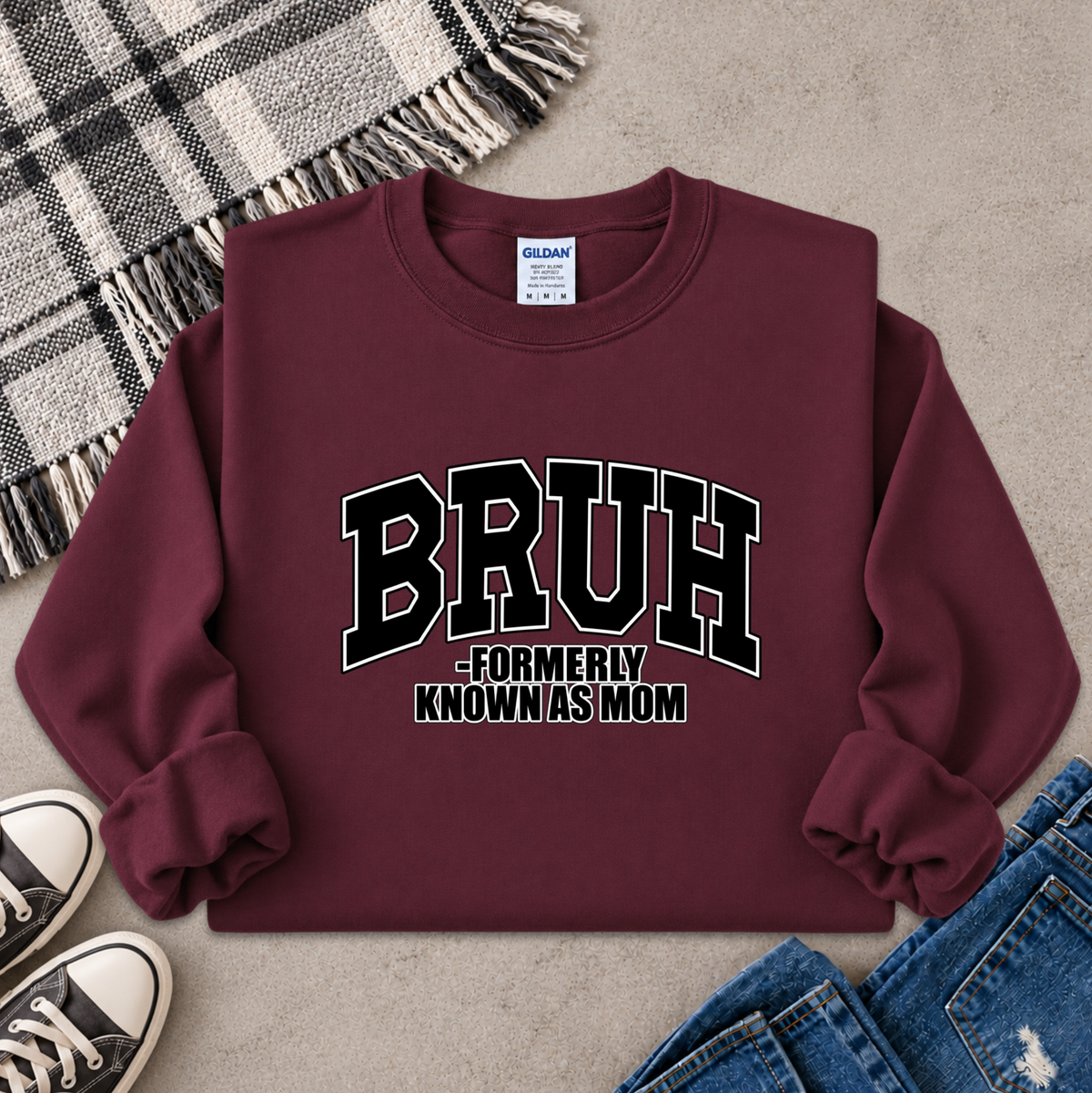 A maroon crewneck sweatshirt with 'BRUH -FORMERLY KNOWN AS MOM' printed in black and white varsity lettering, styled in a flat lay with jeans, sneakers, a plaid blanket, and a gray textured background.