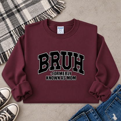 A maroon crewneck sweatshirt with 'BRUH -FORMERLY KNOWN AS MOM' printed in black and white varsity lettering, styled in a flat lay with jeans, sneakers, a plaid blanket, and a gray textured background.