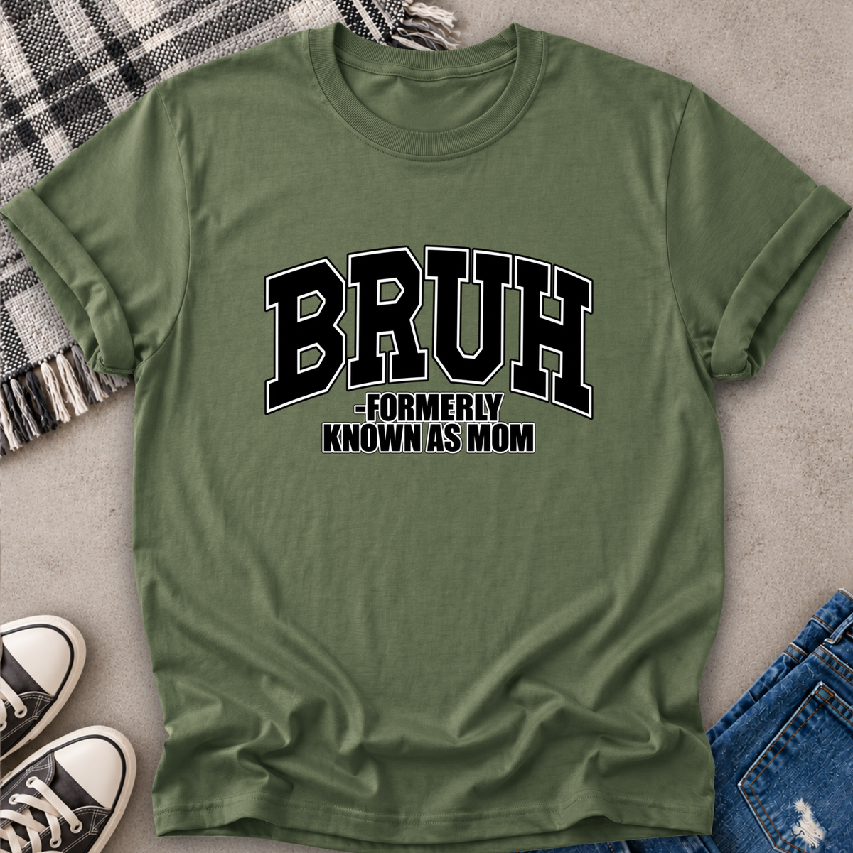 An olive green short-sleeve t-shirt with a black and white graphic reading 'BRUH -FORMERLY KNOWN AS MOM', styled in a flat-lay with jeans, sneakers, and a plaid blanket on a gray textured surface.