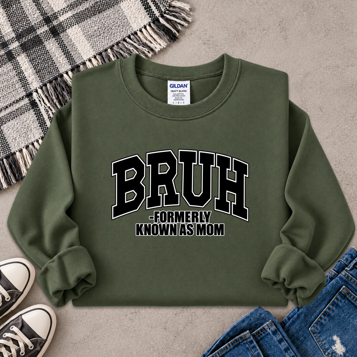An olive green crewneck sweatshirt with 'BRUH -FORMERLY KNOWN AS MOM' printed in black varsity letters with white outlines on the front. The sweatshirt is styled in a flat lay with a plaid blanket, sneakers, and jeans on a gray concrete background.