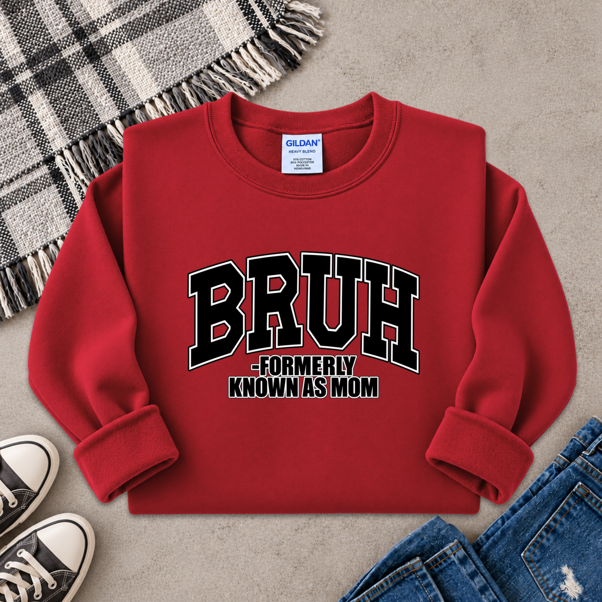 A red long-sleeve sweatshirt laid flat on a textured gray surface, styled with a plaid blanket, sneakers, and jeans. The sweatshirt has a large black and white graphic reading 'BRUH -FORMERLY KNOWN AS MOM' on the front.