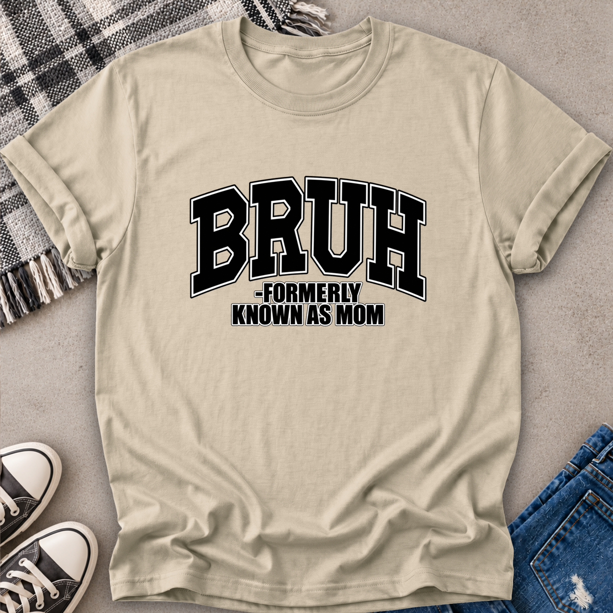 A beige short-sleeve t-shirt with a black graphic text reading 'BRUH -FORMERLY KNOWN AS MOM', styled in a flat-lay with jeans, sneakers, and a plaid blanket on a gray surface.