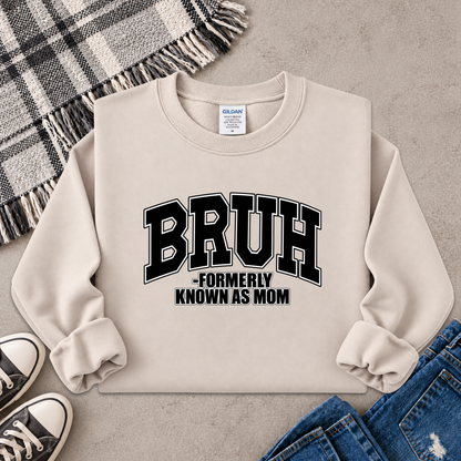 A beige crewneck sweatshirt with 'BRUH -FORMERLY KNOWN AS MOM' printed in black varsity-style lettering on the front. The sweatshirt is styled in a flat lay with a plaid throw blanket, black sneakers, and blue jeans on a gray concrete surface.