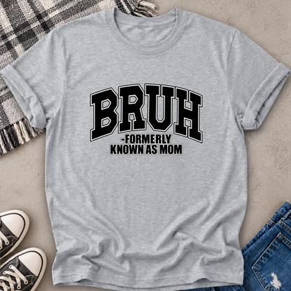 A heather gray short-sleeve t-shirt with a black graphic text reading 'BRUH -FORMERLY KNOWN AS MOM' laid flat on a neutral surface with jeans, sneakers, and a plaid blanket partially visible around it.