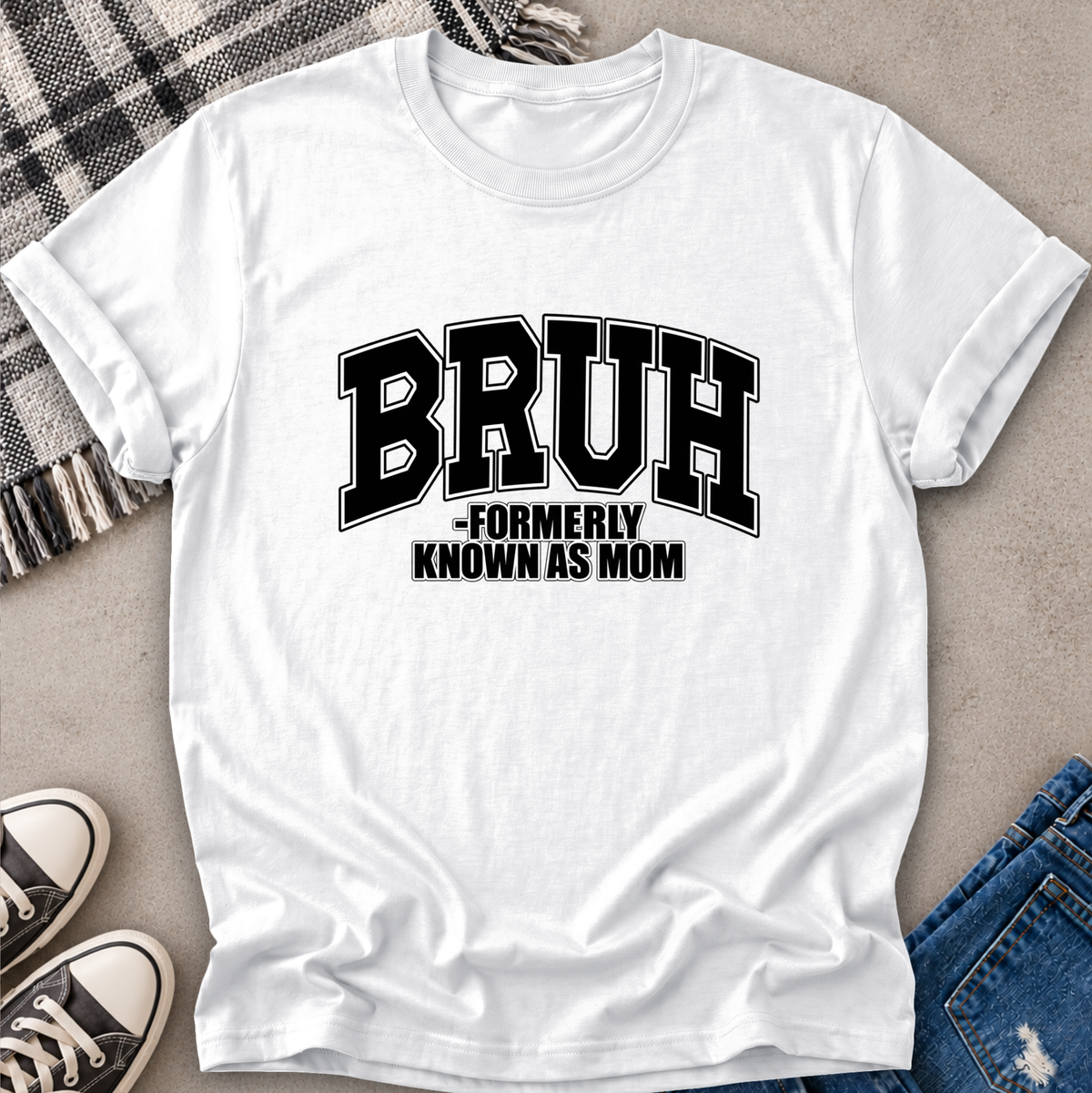 A white short-sleeve T-shirt laid flat on a light gray surface, styled with a plaid blanket, black sneakers, and blue jeans. The shirt has large black collegiate-style text reading “BRUH” with smaller text underneath reading “FORMERLY KNOWN AS MOM.
