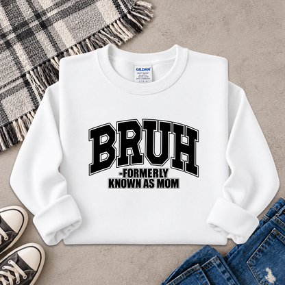 A white long-sleeve sweatshirt laid flat on a textured surface, featuring the text 'BRUH -FORMERLY KNOWN AS MOM' in black varsity-style lettering. A Gildan neck label is visible, and the sweatshirt is styled with a plaid blanket, sneakers, and jeans.