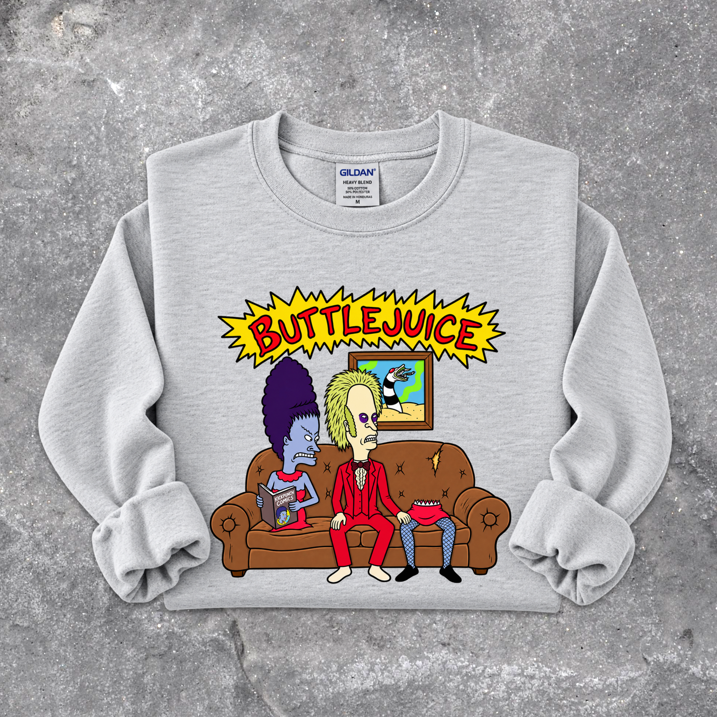 A light gray crewneck sweatshirt with rolled cuffs, featuring a colorful graphic print of two cartoon characters on a brown couch under a yellow 'BUTTLEJUICE' starburst. The sweatshirt is laid flat on a textured gray concrete surface.