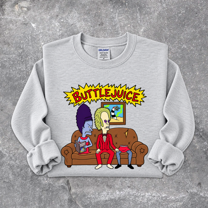 A light gray crewneck sweatshirt with rolled cuffs, featuring a colorful graphic print of two cartoon characters on a brown couch under a yellow 'BUTTLEJUICE' starburst. The sweatshirt is laid flat on a textured gray concrete surface.