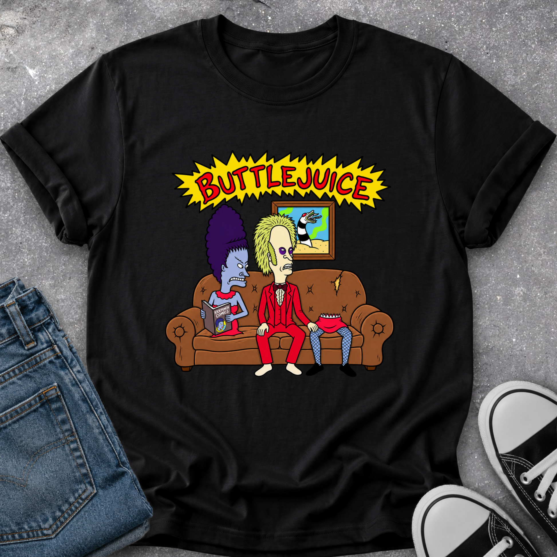 A black graphic t-shirt featuring a cartoon illustration of two characters on a brown couch under a yellow 'BUTTLJUICE' comic-style banner, styled flat-lay on a gray textured surface with blue jeans and black-and-white sneakers partially visible.