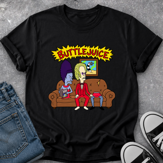 A black graphic t-shirt featuring a cartoon illustration of two characters on a brown couch under a yellow 'BUTTLJUICE' comic-style banner, styled flat-lay on a gray textured surface with blue jeans and black-and-white sneakers partially visible.