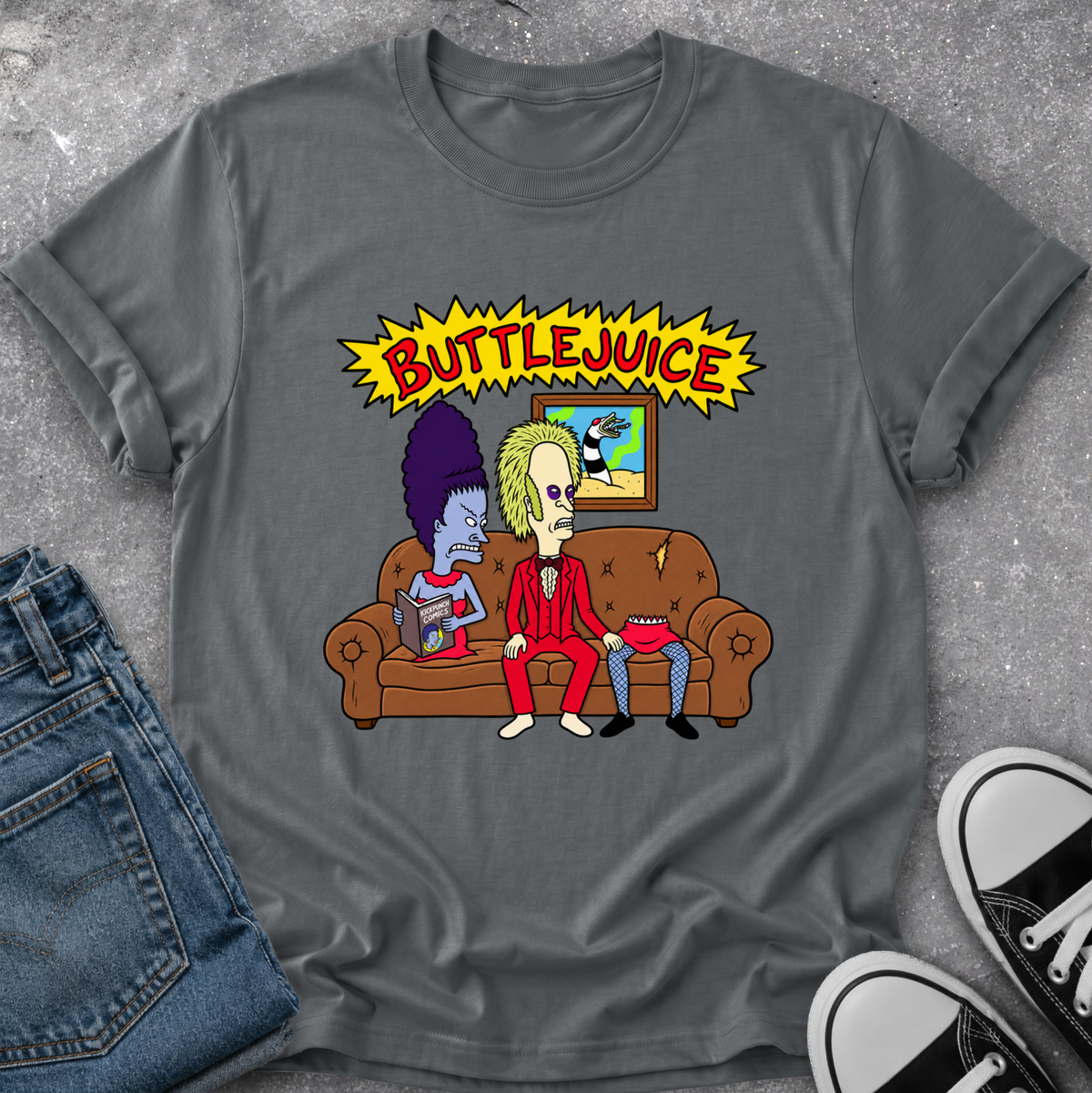 A dark gray graphic t-shirt featuring a cartoon illustration of two characters on a brown couch under a yellow 'BUTTEJUICE' starburst, styled in a flat lay with blue jeans and black-and-white sneakers on a gray textured surface.