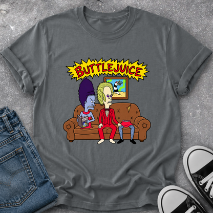 A dark gray graphic t-shirt featuring a cartoon illustration of two characters on a brown couch under a yellow 'BUTTEJUICE' starburst, styled in a flat lay with blue jeans and black-and-white sneakers on a gray textured surface.