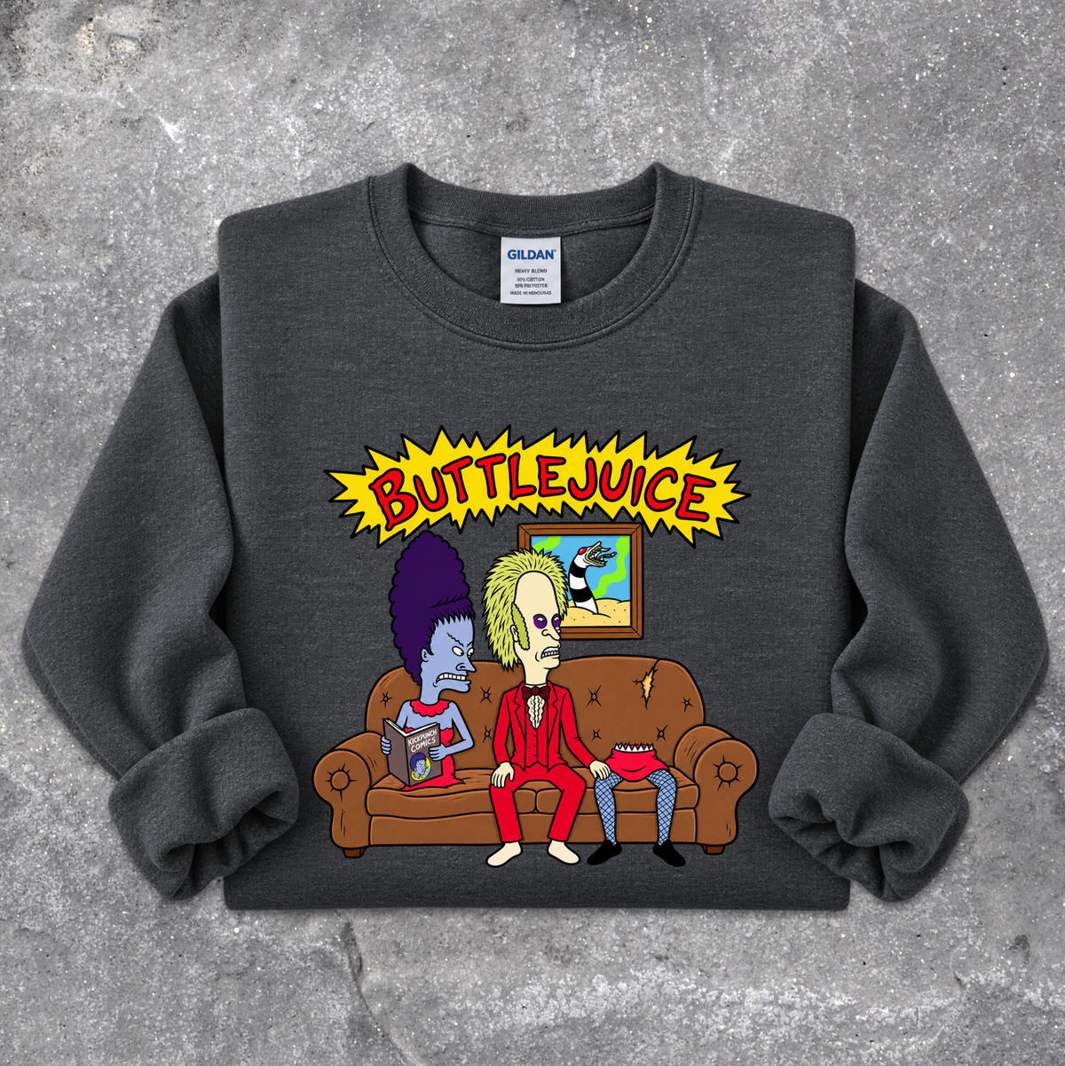 A dark gray crewneck sweatshirt featuring a cartoon graphic of two characters sitting on a brown couch under a yellow 'BUTTLEJUICE' starburst graphic, laid flat on a textured gray concrete background. The neck label shows the brand Gildan.