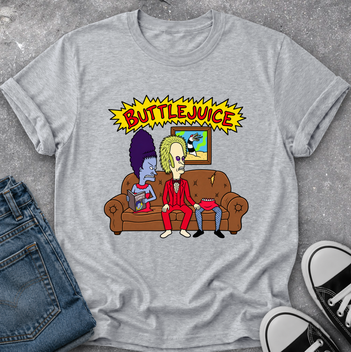 A heather gray graphic t-shirt featuring a cartoon illustration of two characters sitting on a brown couch under a yellow 'BUTTLEJUICE' comic-style banner. The shirt is styled in a flat lay with blue jeans and black-and-white sneakers on a gray textured surface.