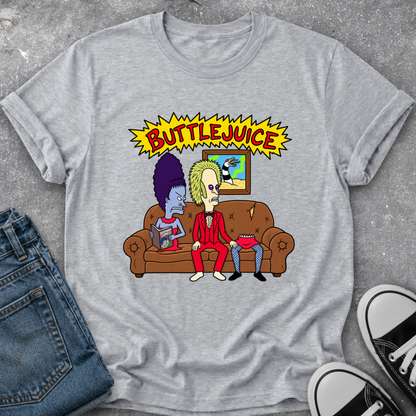 A heather gray graphic t-shirt featuring a cartoon illustration of two characters sitting on a brown couch under a yellow 'BUTTLEJUICE' comic-style banner. The shirt is styled in a flat lay with blue jeans and black-and-white sneakers on a gray textured surface.