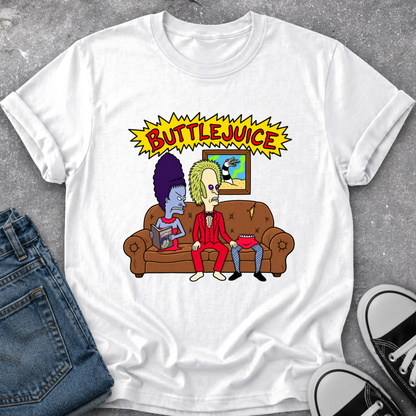 A white graphic t-shirt featuring a cartoon illustration of two characters sitting on a brown couch under a yellow 'BUTTLEJUICE' comic-style banner. The shirt is styled in a flat lay with blue jeans and black-and-white sneakers on a gray textured background.