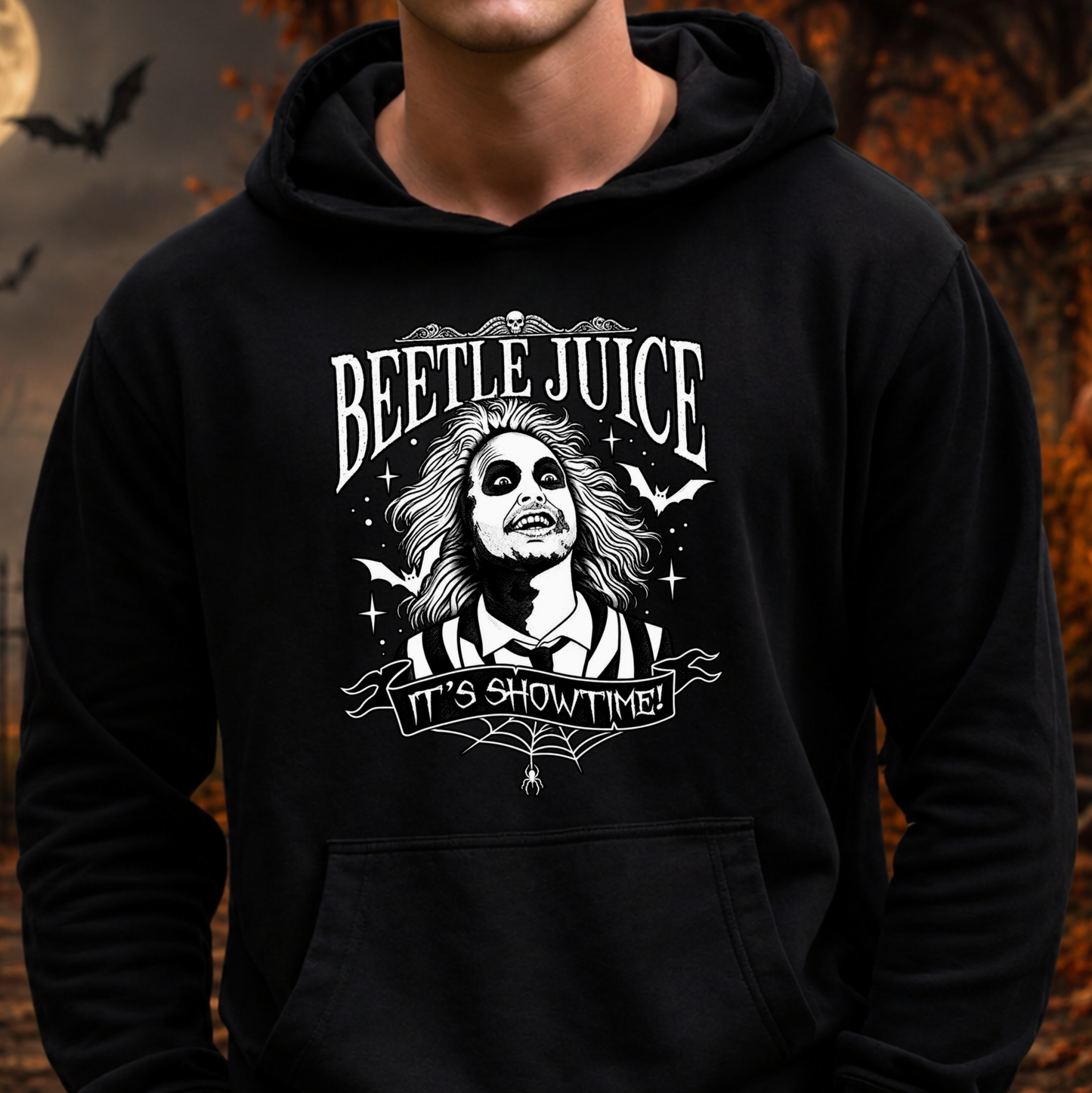 A person wearing a black pullover hoodie with a large white Beetlejuice graphic on the chest that reads 'BEETLE JUICE' and 'IT'S SHOWTIME!'. The background is a blurred Halloween-themed outdoor scene with bats and autumn leaves.