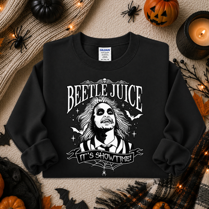 A black crewneck sweatshirt with a white graphic of Beetlejuice and the text 'BEETLE JUICE' and 'IT'S SHOWTIME!' laid flat on a cozy autumn-themed background with pumpkins, a candle, and spider decorations.
