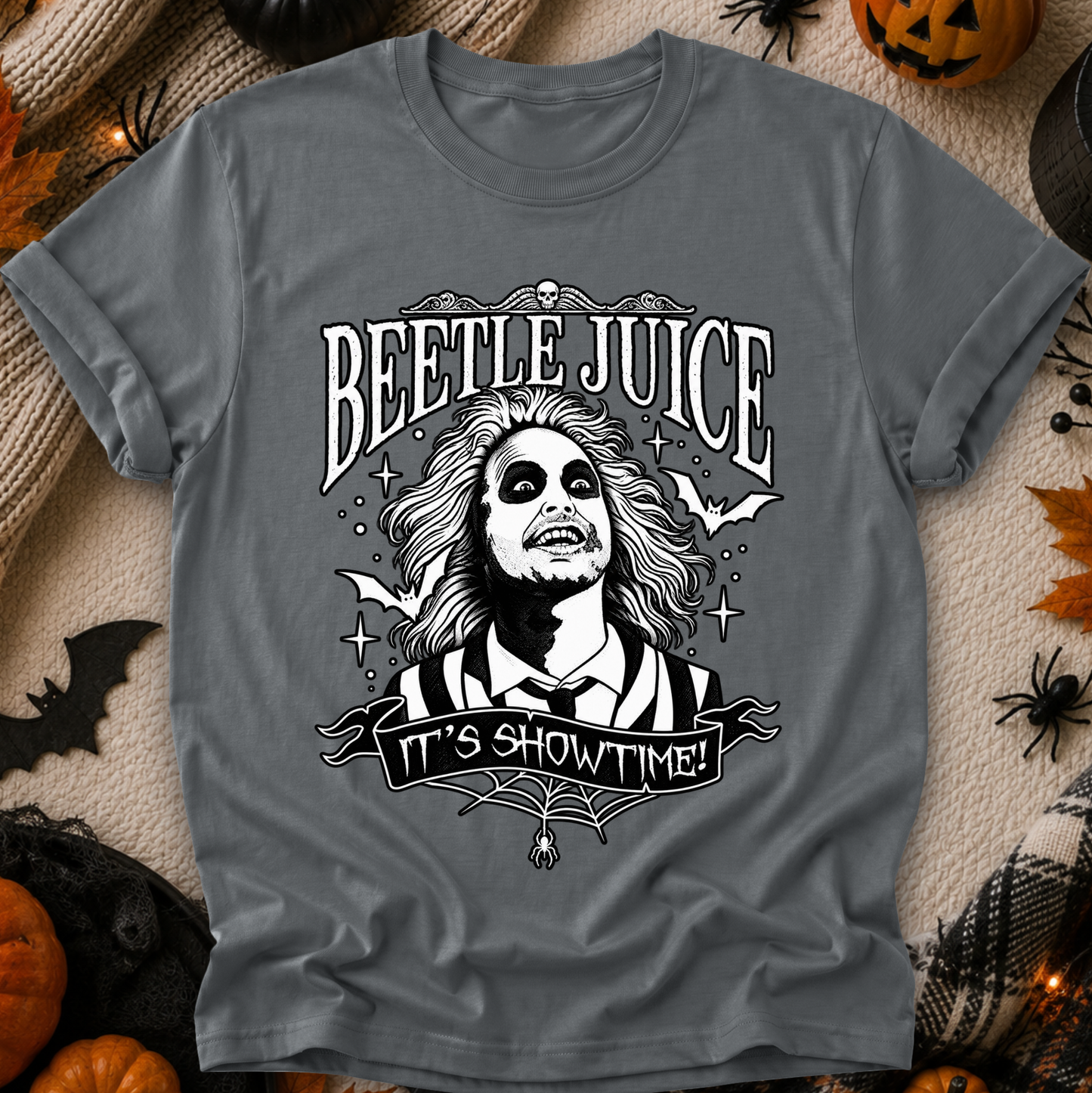A gray short-sleeve graphic T-shirt laid flat on a textured beige fabric with Halloween props around it. The shirt features a black-and-white Beetlejuice illustration with the words “BEETLE JUICE” and “IT’S SHOWTIME!”.