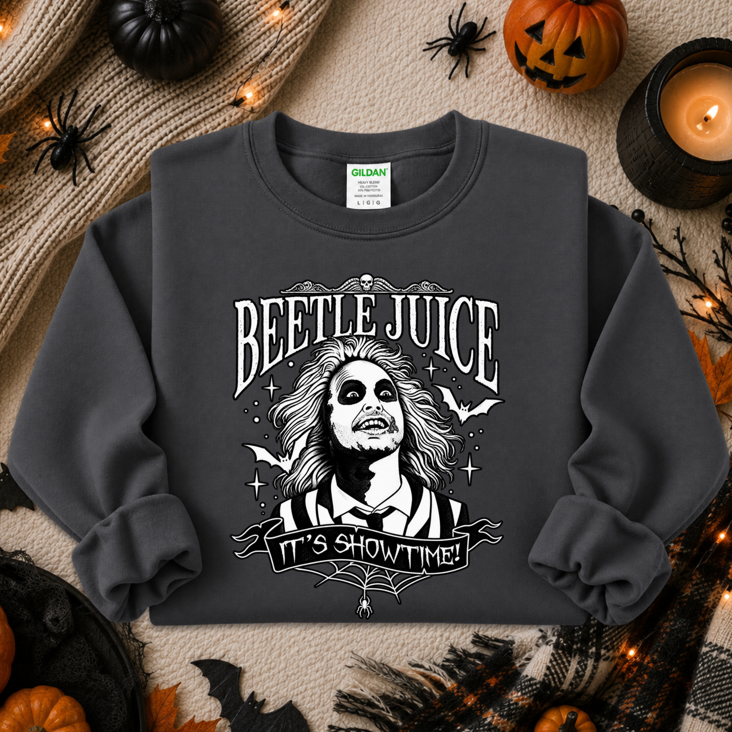 A dark charcoal gray crewneck sweatshirt featuring a white graphic of Beetlejuice with the text 'BEETLE JUICE' and 'IT'S SHOWTIME!'. The sweatshirt is displayed in a flat-lay Halloween setting with pumpkins, a candle, and spider decorations.