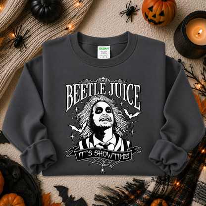 A dark charcoal gray crewneck sweatshirt featuring a white graphic of Beetlejuice with the text 'BEETLE JUICE' and 'IT'S SHOWTIME!'. The sweatshirt is displayed in a flat-lay Halloween setting with pumpkins, a candle, and spider decorations.