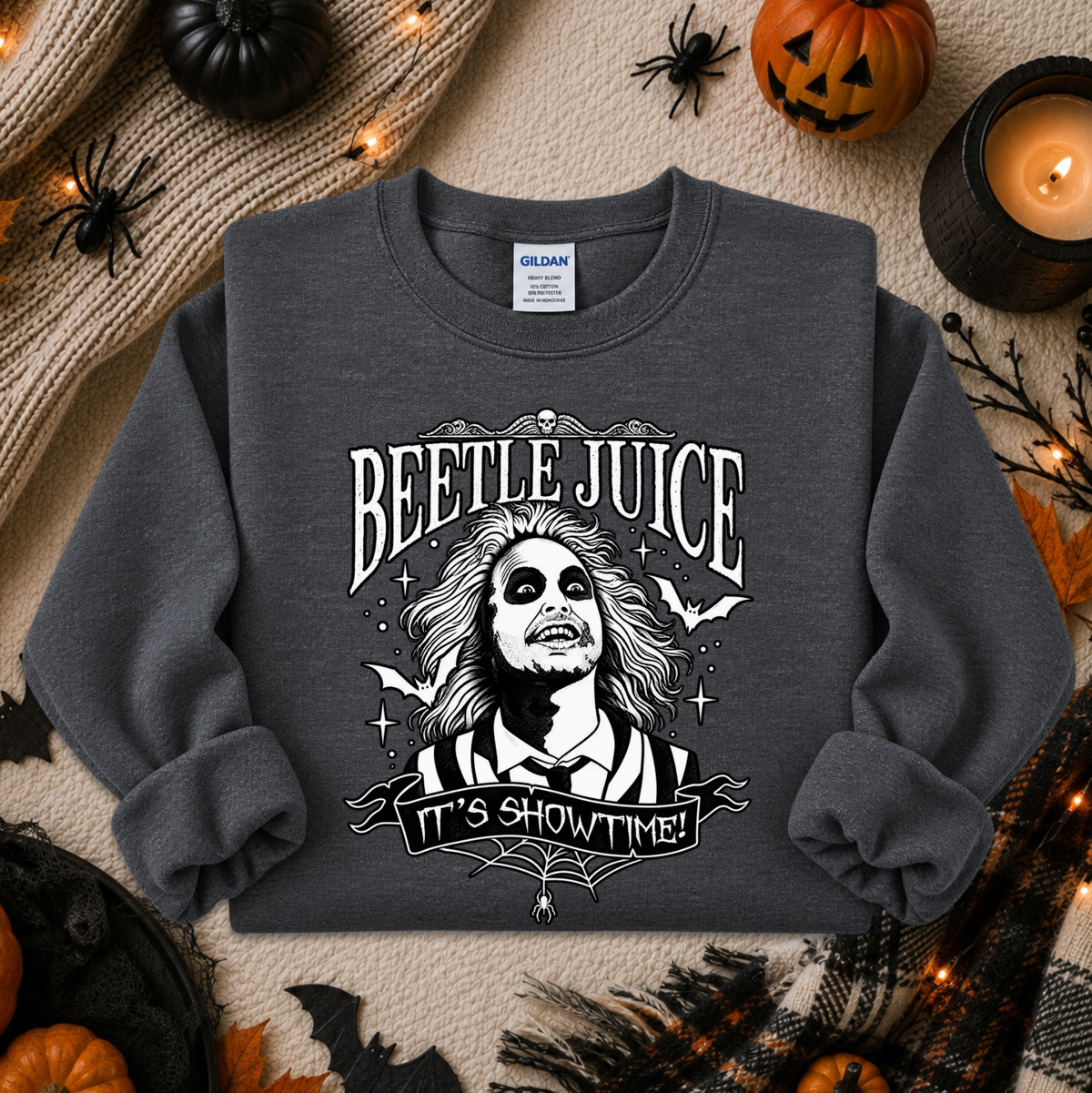 A dark gray crewneck sweatshirt featuring a black and white graphic of Beetlejuice with the text 'BEETLE JUICE' and 'IT'S SHOWTIME!'. The sweatshirt is displayed in a flat-lay Halloween setting with pumpkins, a candle, and spider decorations.