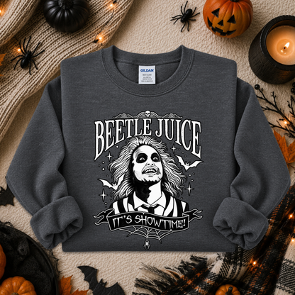 A dark gray crewneck sweatshirt featuring a black and white graphic of Beetlejuice with the text 'BEETLE JUICE' and 'IT'S SHOWTIME!'. The sweatshirt is displayed in a flat-lay Halloween setting with pumpkins, a candle, and spider decorations.