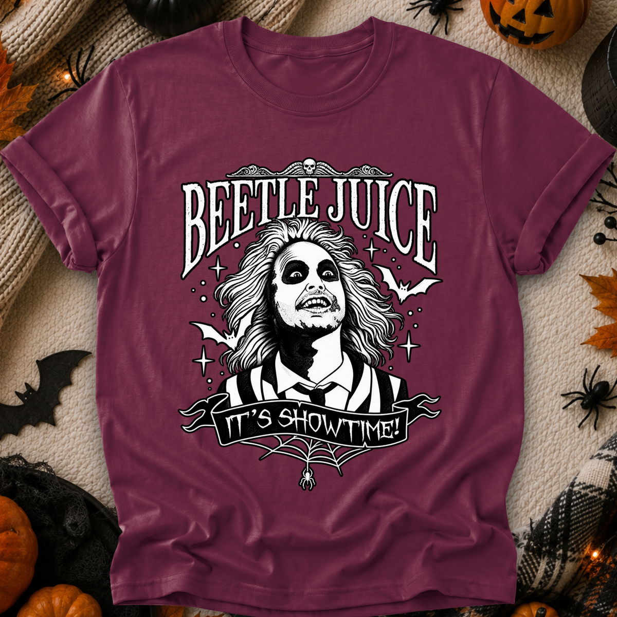 A maroon short-sleeve graphic T-shirt laid flat on a Halloween-themed background with pumpkins, spider decorations, and autumn leaves. The shirt features a large black-and-white Beetlejuice portrait and the words “BEETLE JUICE” and “IT’S SHOWTIME!” printed on the front.