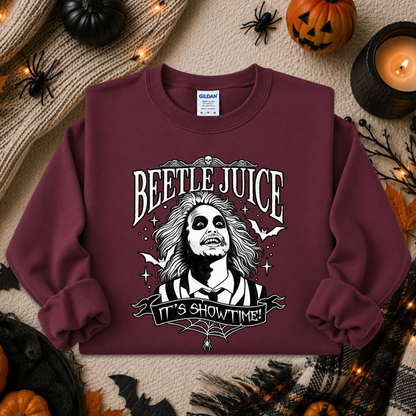 A maroon crewneck sweatshirt with a 'Beetlejuice' graphic print, styled in a flat lay with Halloween props like pumpkins, a candle, and spider decorations on a beige knit blanket.
