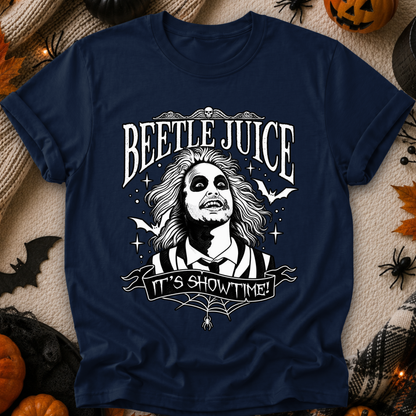 A navy blue graphic t-shirt featuring Beetlejuice with the text 'BEETLE JUICE' and 'IT'S SHOWTIME!' in white. The shirt is displayed in a flat-lay Halloween setting with pumpkins, spider decorations, and a beige knit blanket.