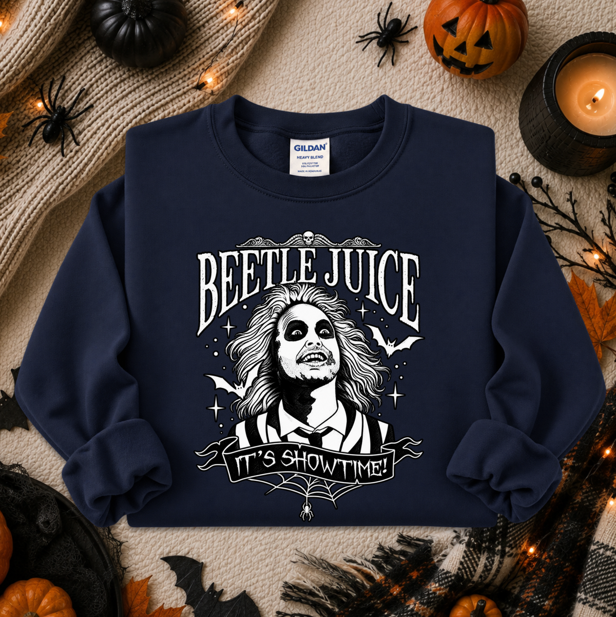 A navy blue crewneck sweatshirt with a white graphic of Beetlejuice and the text 'BEETLE JUICE' and 'IT'S SHOWTIME!' laid flat on a Halloween-themed background with pumpkins, a candle, and spider decorations.