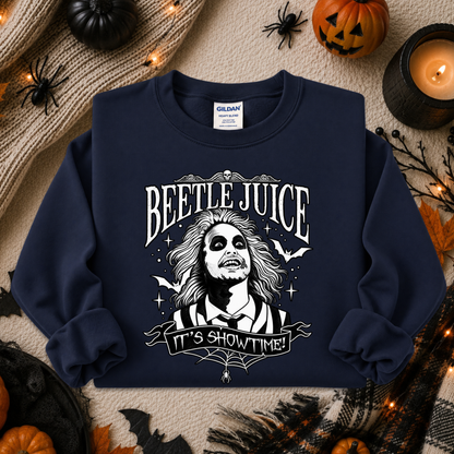 A navy blue crewneck sweatshirt with a white graphic of Beetlejuice and the text 'BEETLE JUICE' and 'IT'S SHOWTIME!' laid flat on a Halloween-themed background with pumpkins, a candle, and spider decorations.