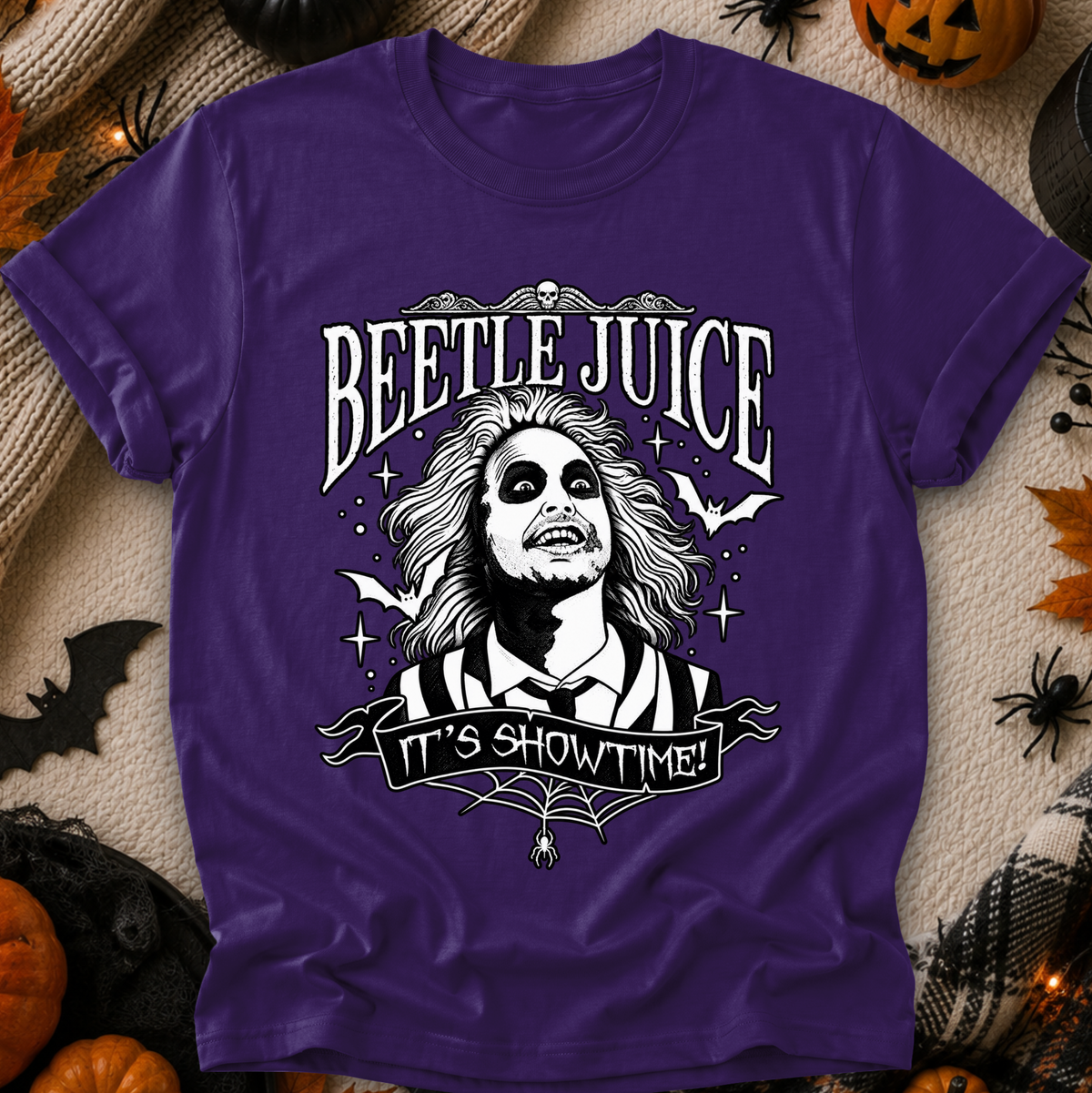 A purple short-sleeve graphic T-shirt laid flat on a Halloween-themed background with pumpkins, spider decorations, and a blanket. The shirt features a large white illustrated character and the text 'BEETLE JUICE' and 'IT'S SHOWTIME!'.