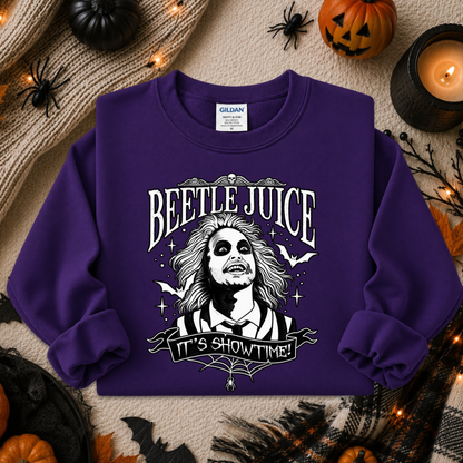 A purple crewneck sweatshirt with a 'Beetle Juice It's Showtime!' graphic, styled in a flat lay with Halloween props including pumpkins, a candle, and spider decorations on a beige knit blanket.