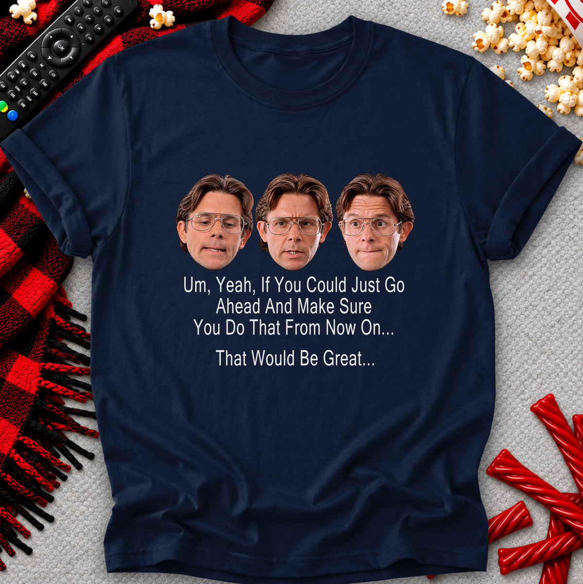 A navy blue graphic t-shirt laid flat on a gray textured background with holiday-themed props like a remote control, popcorn, and red-and-black plaid fabric. The shirt features a humorous quote in white text below three repeated faces of a man with glasses.