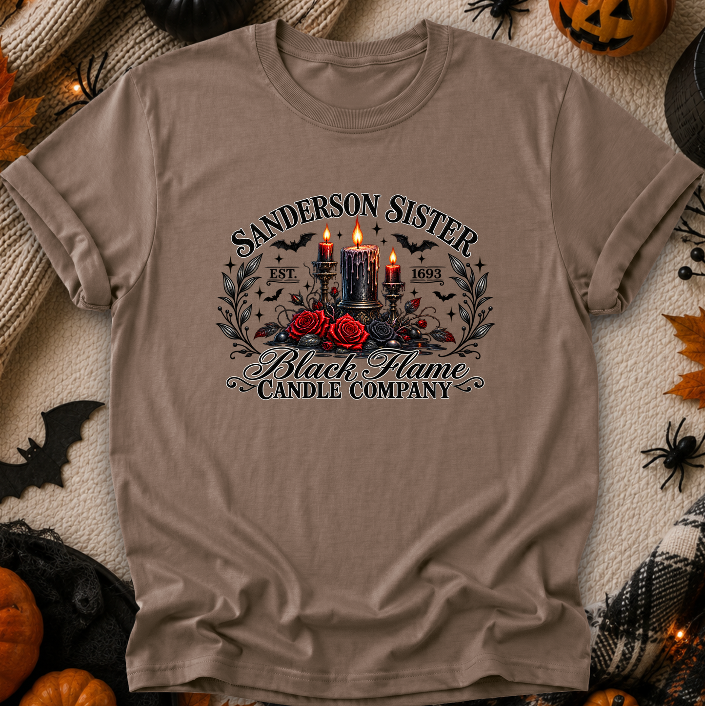 A taupe brown graphic t-shirt laid flat on a cozy, Halloween-themed background. The shirt features a gothic-style graphic with the text 'SANDERSON SISTER' and 'Black Flame Candle Company' surrounding an illustration of two burning black candles, red roses, and bats.
