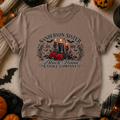 A taupe brown graphic t-shirt laid flat on a cozy, Halloween-themed background. The shirt features a gothic-style graphic with the text 'SANDERSON SISTER' and 'Black Flame Candle Company' surrounding an illustration of two burning black candles, red roses, and bats.