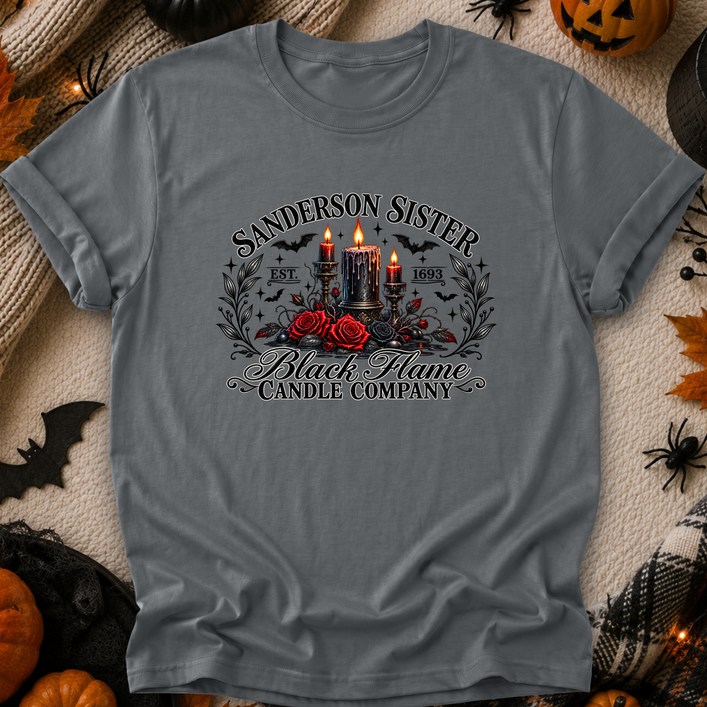 A gray short-sleeve graphic T-shirt laid flat on a cozy beige fabric background with Halloween decorations around it. The shirt features a gothic-style front print reading 'Sanderson Sister Black Flame Candle Company' with three lit black candles, red roses, bats, and decorative text.