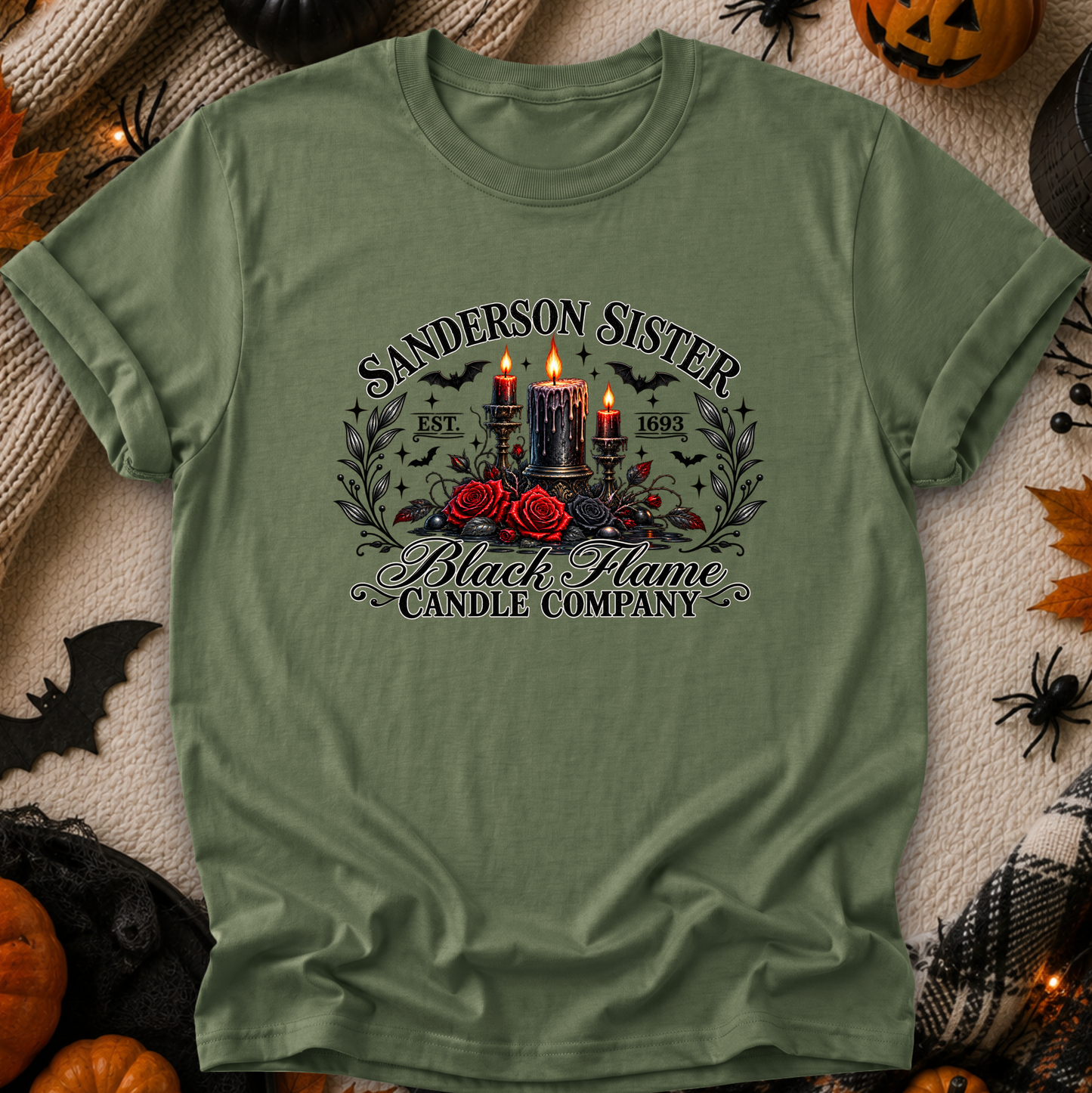 An olive green graphic t-shirt laid flat on a cozy, Halloween-themed background. The shirt features a gothic-style graphic with the text 'SANDERSON SISTER' and 'Black Flame Candle Company' surrounding an illustration of three lit black candles, red roses, and bats.